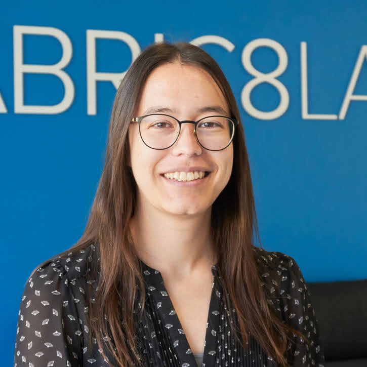 A woman with round glasses, straight brown shoulder length hair, and a black blouse sitting in front of a bright blue wall.