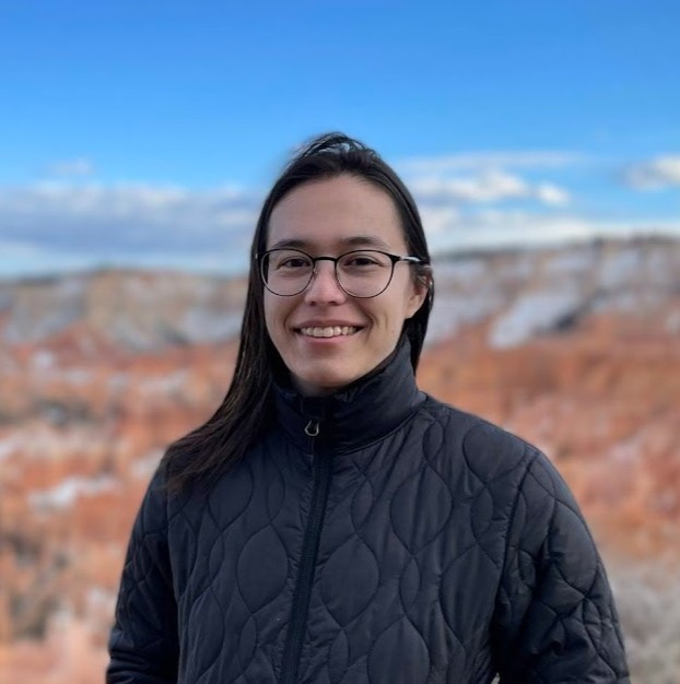 A woman with round glasses, straight brown shoulder length hair, and a black winter jacket standing in front of the jagged orange hoodoos at Bryce Canyon National Park.